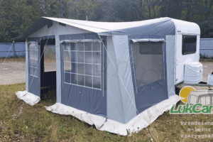 Duży, nowoczesny przedsionek przymocowany do białej przyczepy kempingowej, rozstawiony na zielonym trawniku. Przedsionek wykonany z trwałego materiału w odcieniach szarości, z dużymi oknami po bokach, zapewniającymi dostęp światła dziennego. Na przedsionku widoczne logo "LukCamp.pl". W tle widać dom oraz ogród.