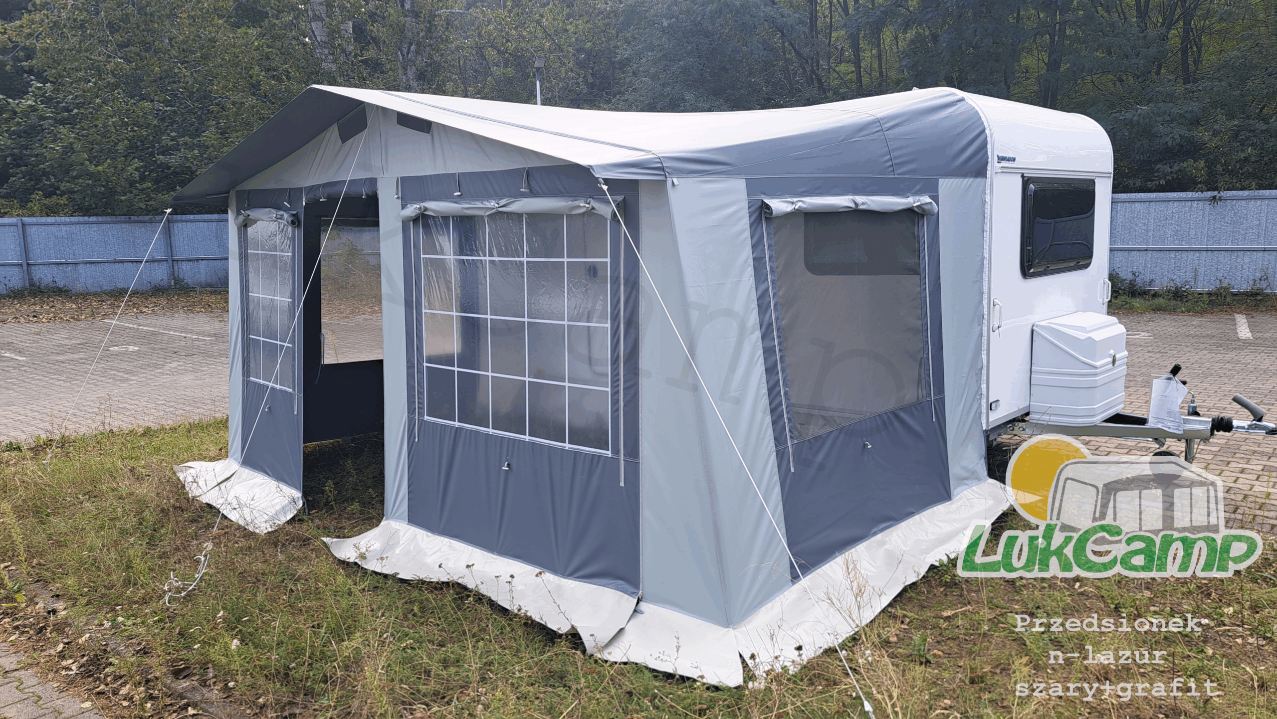 Duży, nowoczesny przedsionek przymocowany do białej przyczepy kempingowej, rozstawiony na zielonym trawniku. Przedsionek wykonany z trwałego materiału w odcieniach szarości, z dużymi oknami po bokach, zapewniającymi dostęp światła dziennego. Na przedsionku widoczne logo "LukCamp.pl". W tle widać dom oraz ogród.
